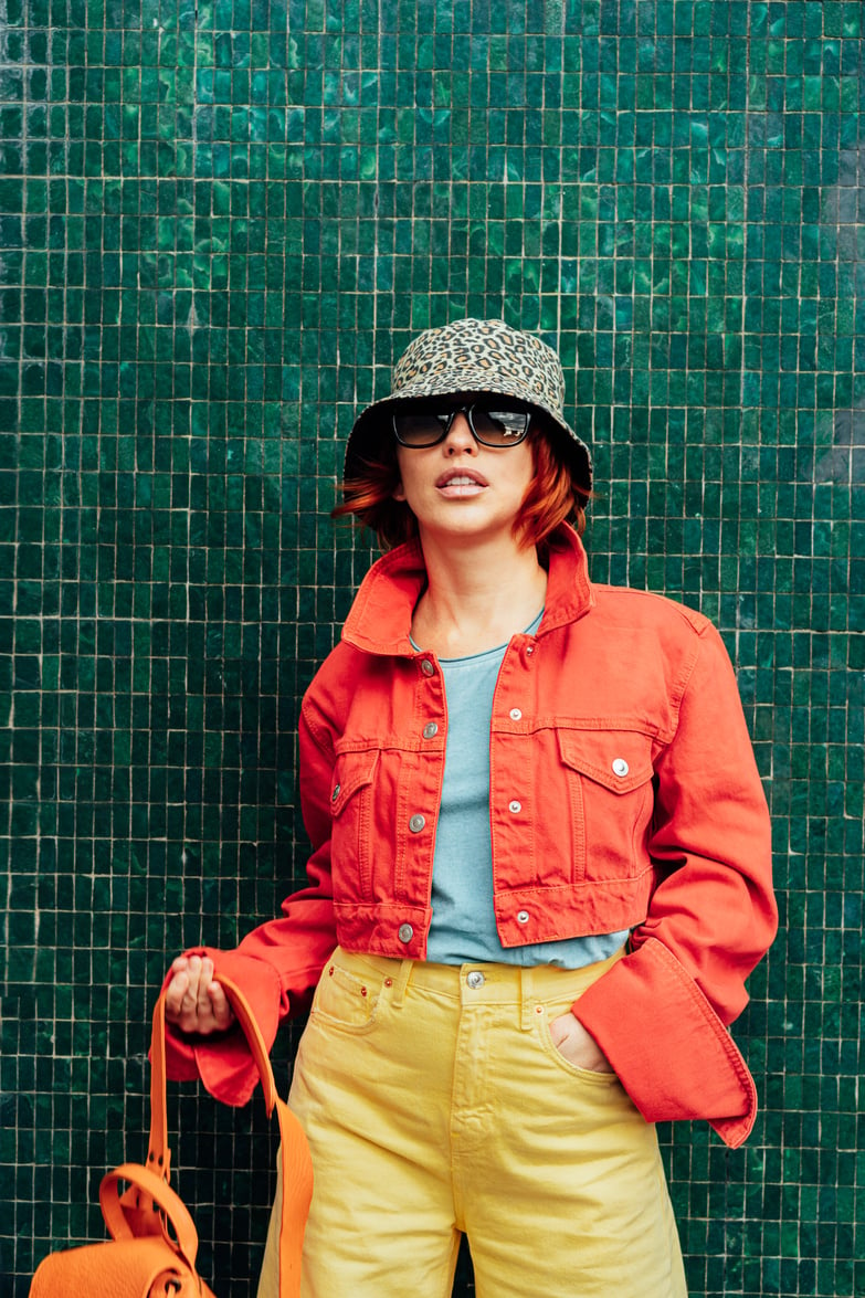 Hipster Fashion Young Woman in Bright Clothes, Sun Glasses and Bucket Hat Posing on the Green Tile Wall Background. Urban City Street Fashion. Vertical Card. Selective Focus. Copy Space